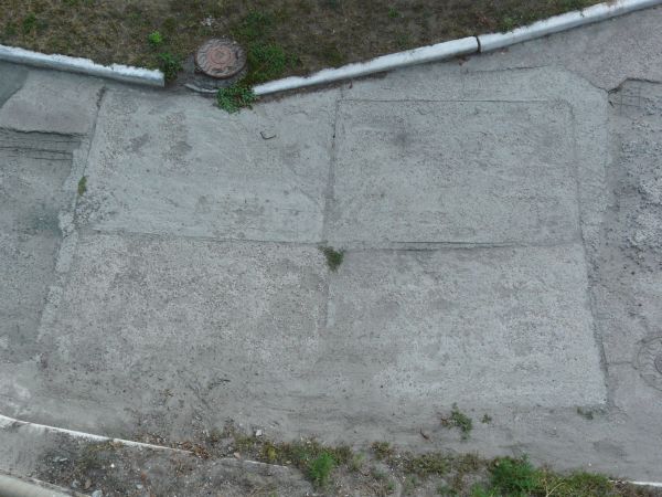 Rough grey asphalt texture, formed into a path between patches of green grass and rocky, sandy weeds.